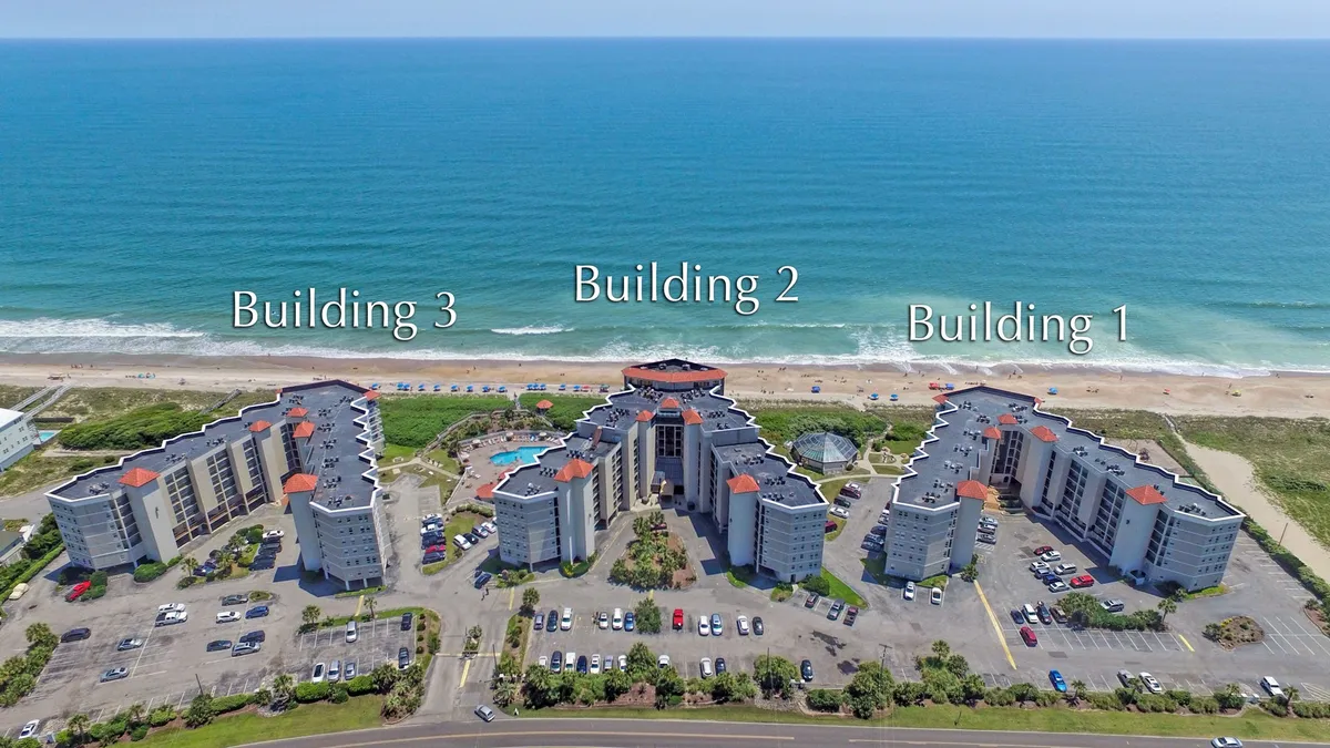 An expansive shot capturing three buildings surrounding a spacious swimming pool just steps away from the sandy beach. The pool area is inviting for both relaxation and exercise, with the beach offering further opportunities for outdoor adventures.