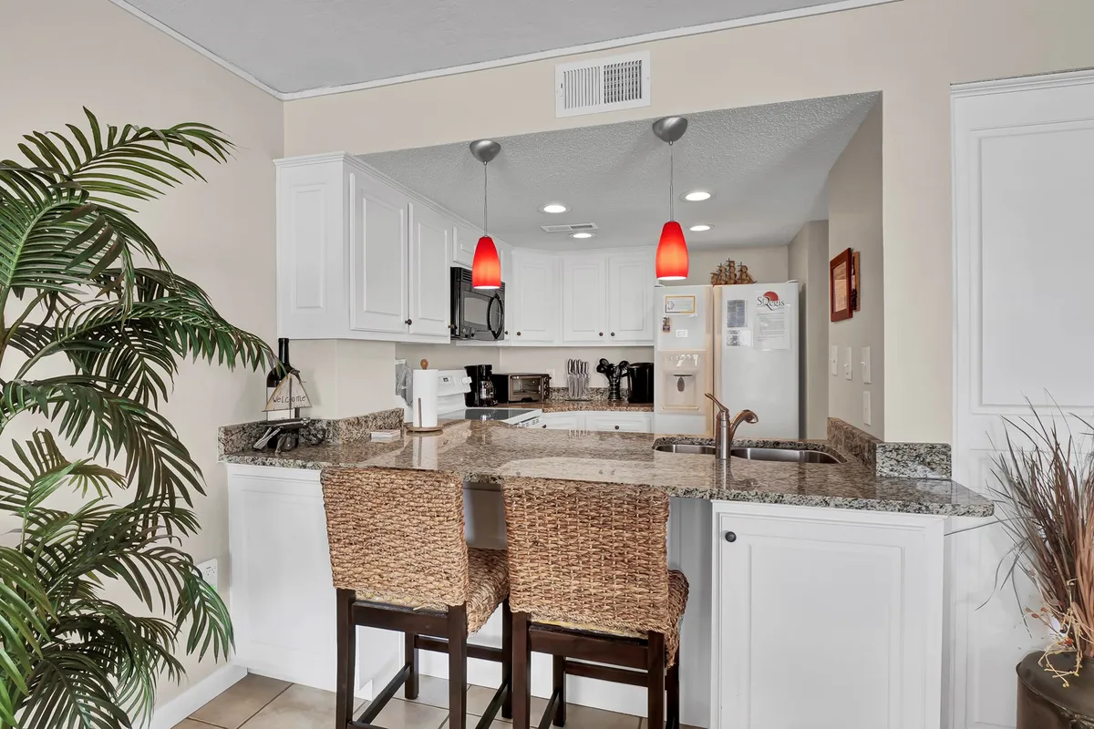 A stylish, inviting kitchen space featuring gleaming granite countertops, bar stools for casual dining, striking red pendant lights, and visible stainless steel appliances. The kitchen layout is open, allowing easy interaction between chef and guests.