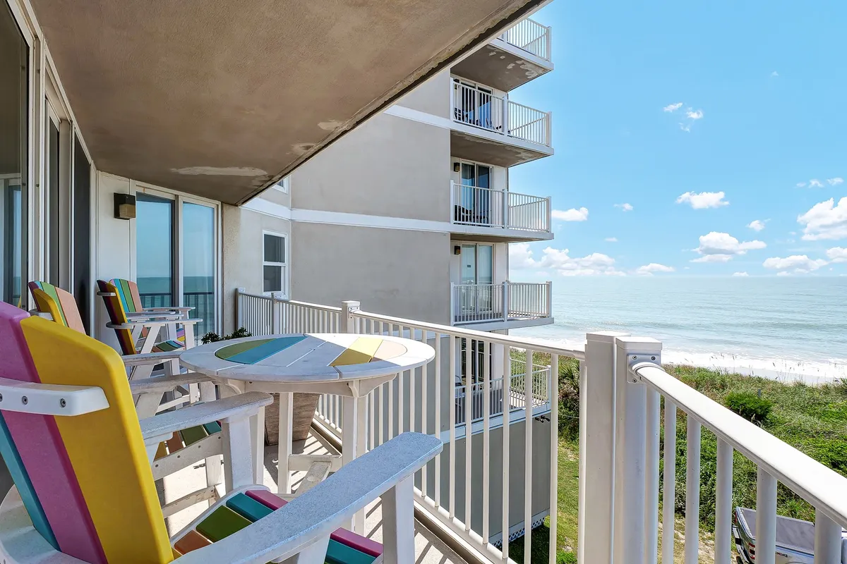 An exterior shot of a patio with colorful chairs arranged around a round table, all set against a stunning ocean backdrop. The scene exudes a cheerful, social atmosphere perfect for relaxation or informal work sessions outside.