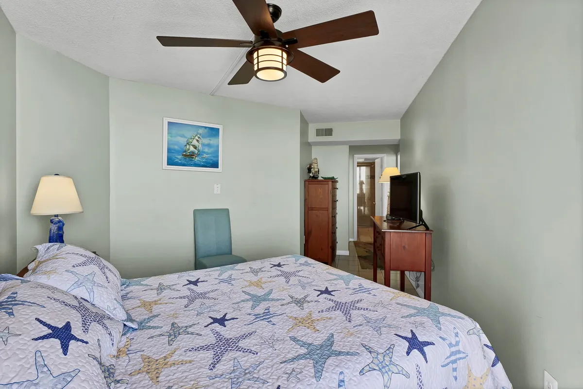 This image features a cozy bedroom with a Queen bed adorned in a starfish-patterned quilt, complemented by a ceiling fan, nautical wall art, a blue accent chair, and a wooden dresser with a TV. The room is bathed in soft, inviting light, creating a relaxing and tranquil atmosphere.