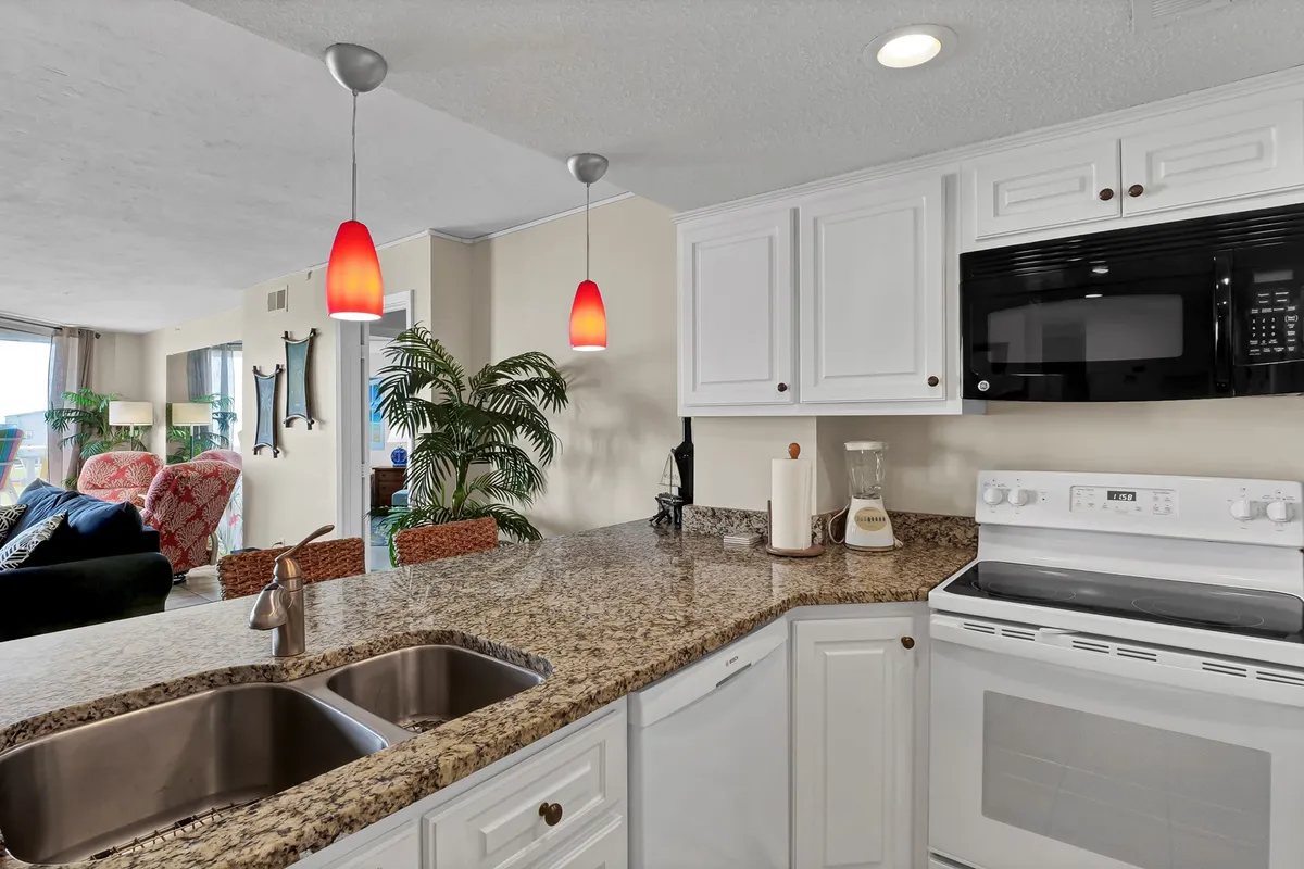 A close-up view of a modern kitchen island with granite countertops, bright red pendant lighting, and sleek white cabinetry. A green plant adds a touch of freshness, and stainless appliances are visible in the background, reinforcing the kitchen's updated style.