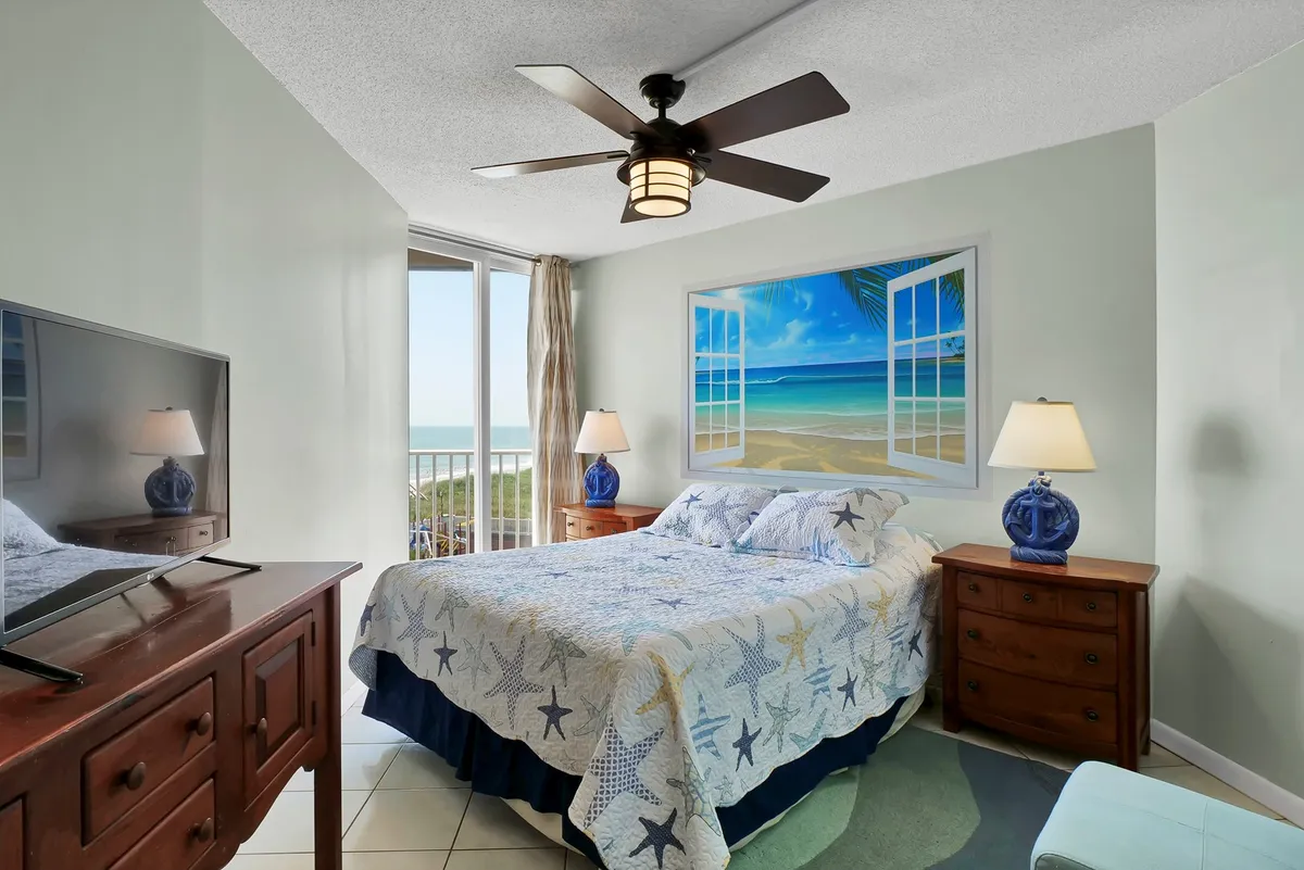 A tranquil bedroom featuring a comfortable Queen bed, a window offering a glimpse of natural light or a beachy view, tasteful wall art, a nightstand, and a television. The setting exudes calm, perfect for unwinding after a day at the shore.