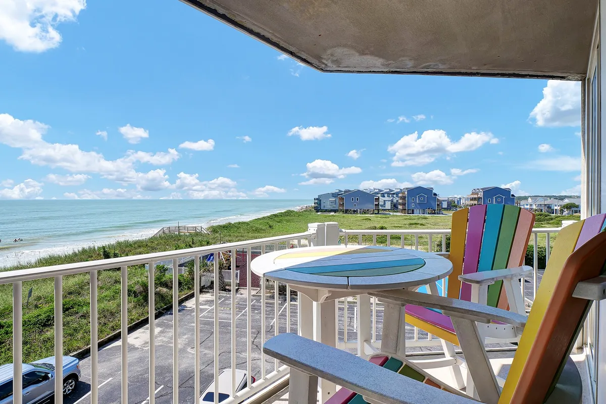 The image showcases a row of vibrant, colorful chairs on a spacious balcony overlooking a breathtaking ocean view under a clear blue sky. The tranquil scene invites guests to relax, enjoy the soothing sea breeze, and soak in the sunrise or unwind with loved ones as the sun sets.