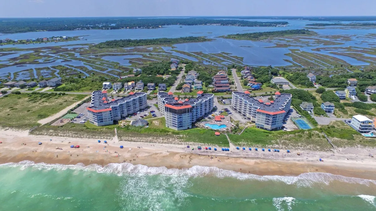 A vibrant scene of a beachfront property with a sparkling swimming pool, colorful umbrellas, and the ocean waves visible beyond. The pool area offers ample space for relaxation or family fun, seamlessly connecting to the sandy coastline.