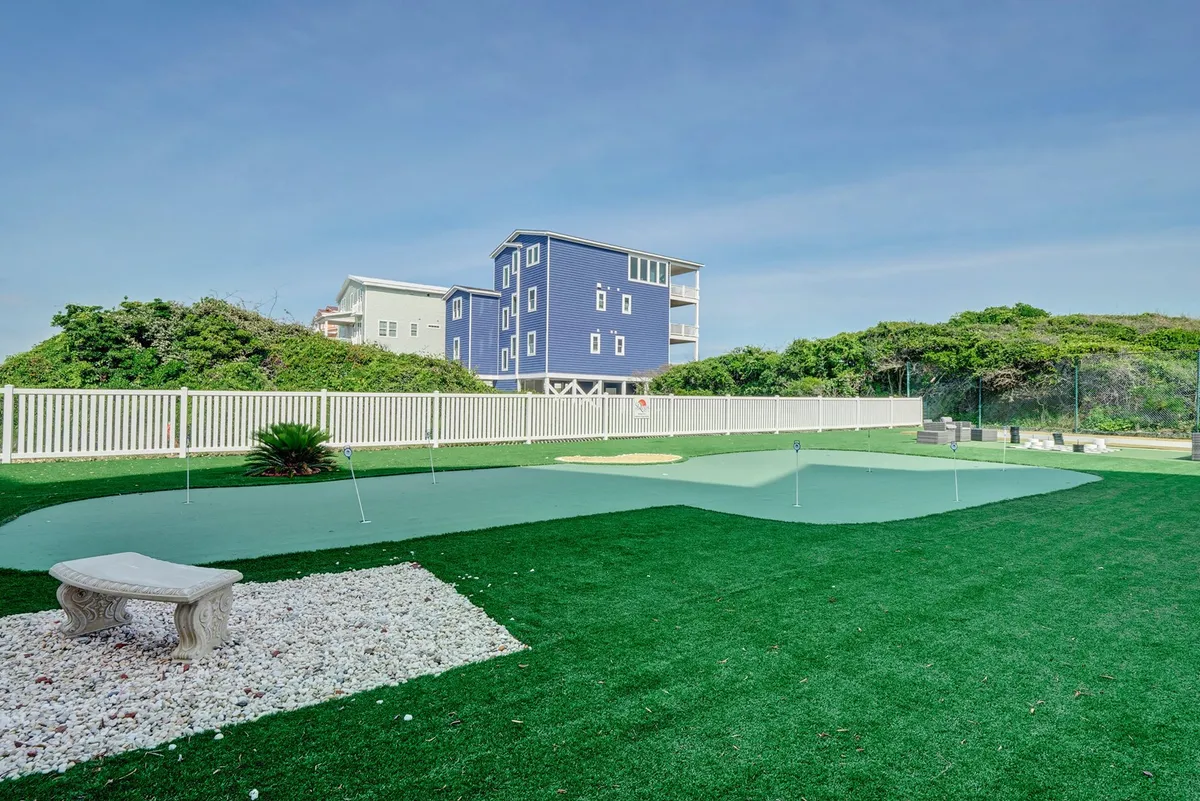A well-maintained synthetic golf putting green bordered by a white picket fence, stone bench, and lush vegetation. The tranquil setting invites guests to practice their swing or enjoy a relaxing afternoon outdoors.