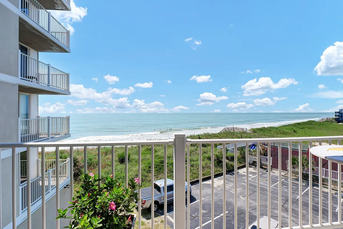 A serene balcony scene overlooking the ocean, with lush green foliage and clouds in the distance. The setting is tranquil, inviting relaxation or a peaceful moment away from work, and showcases the proximity to the beach and beautiful sunrise or sunset views.