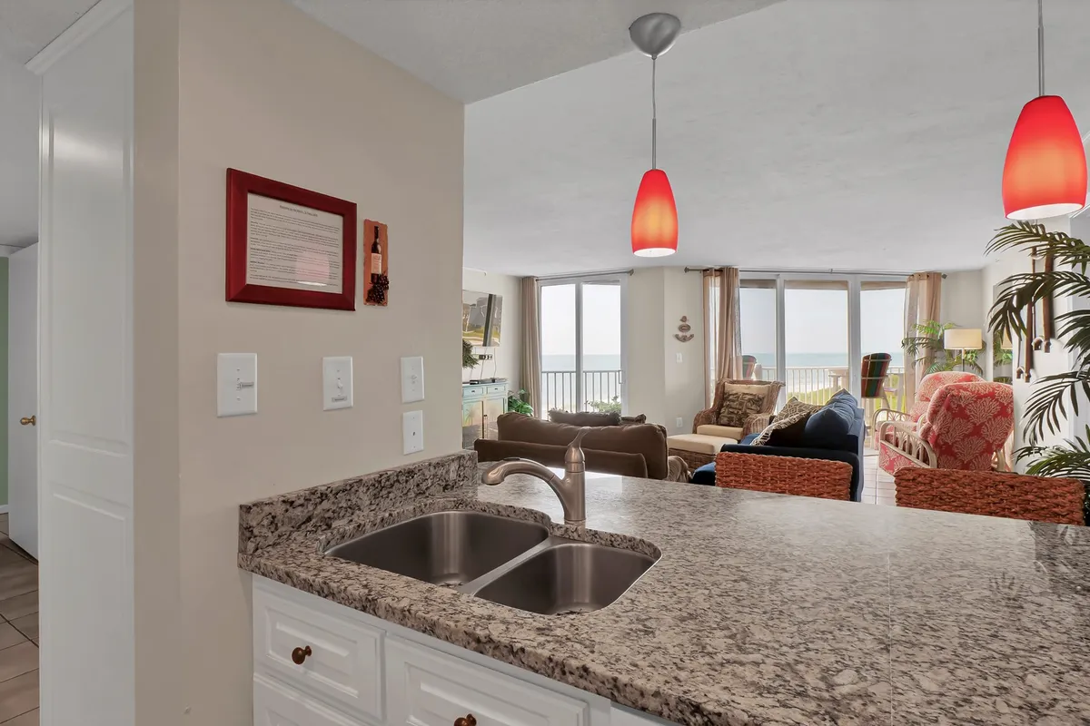 A spacious living area with plush seating, granite countertop, red pendant lights, and expansive windows offering panoramic ocean views. The space is simultaneously cozy and airy, designed for both relaxation and entertaining guests.
